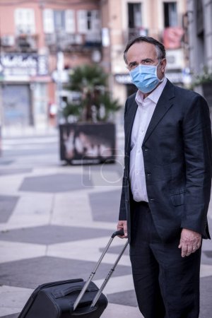 Photo for Tourist dressed in suit with protective face mask and trolley suitcase is in urban context - Royalty Free Image
