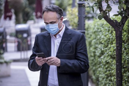 Photo for Man dressed in suit with protective face mask uses his cellphone in an urban context - Royalty Free Image