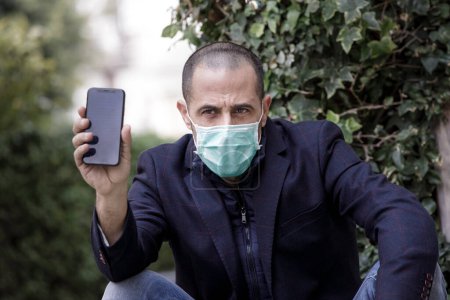 Foto de Hombre afeitado en chaqueta de mascarilla verde sentado en un banco muestra la pantalla de su teléfono celular que dice STOP "Covid-19" - Imagen libre de derechos