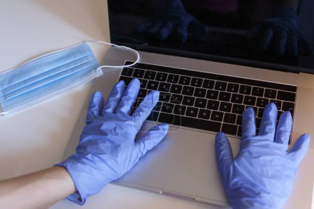 Téléchargez les photos : Mains avec des gants en latex travaillent sur l'ordinateur sur une table blanche avec un masque chirurgical près - en image libre de droit