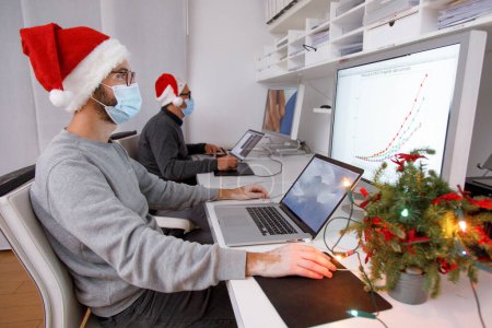 Foto de Hombres con máscaras médicas trabajando en la oficina con un ordenador portátil, concepto de Navidad, año nuevo, pandemia coronavirus - Imagen libre de derechos