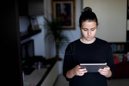 Téléchargez les photos : Adolescent aux cheveux foncés recueilli et une chemise noire regarde la tablette dans le salon - en image libre de droit