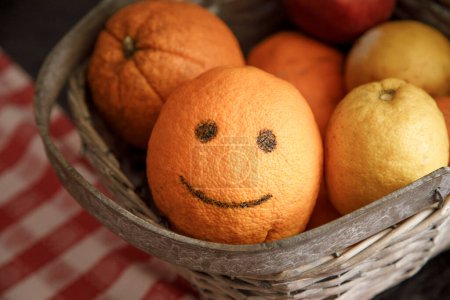 Téléchargez les photos : Panier d'oranges, dont l'un dessiné un sourire à l'intérieur d'un panier en osier sur une table avec nappe à carreaux - en image libre de droit