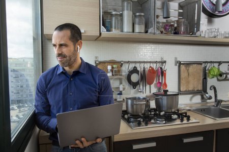 Téléchargez les photos : Homme brun avec chemise bleue travaille à l'ordinateur dans le travail intelligent assis dans la cuisine - en image libre de droit