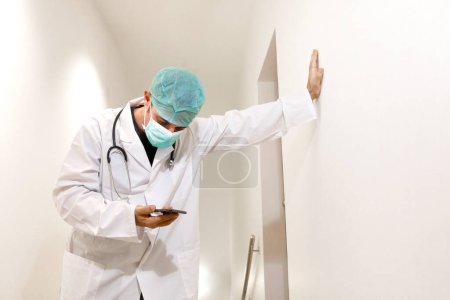 Foto de Médico con máscara y gorra en la sala de un hospital devastado por la fatiga - Imagen libre de derechos