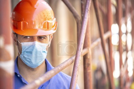 Photo for Engineer, under the scaffolding of a construction site he wears a protective helmet and a surgical mask - Royalty Free Image