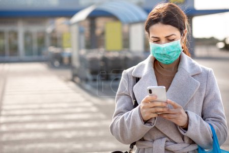Photo for Girl with coat and facial surgical mask, stops in front of the supermarket with the shopping bag and speaks on a cell phone - Royalty Free Image