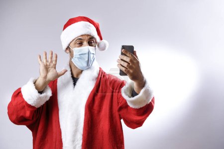 Foto de Santa Claus con máscara quirúrgica utiliza su celular para hacer una videollamada, aislada sobre fondo blanco - Imagen libre de derechos