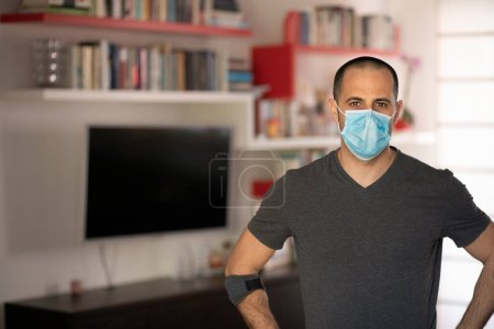 Téléchargez les photos : Rasé homme brun avec chemise grise et masque chirurgical, se tient dans le salon à la maison - en image libre de droit