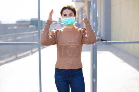 Photo for Brunette girl with orange sweater adjusts her facial mask, isolated on wire mesh. - Royalty Free Image
