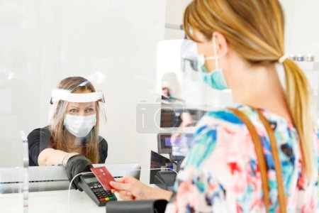 Photo for Blonde girl with mask pays at the checkout with a credit card where the cashier is protected by a plexiglass plate and a visor with mask - Royalty Free Image