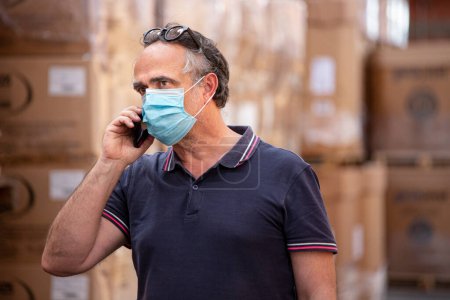 Photo for Businessman with surgical mask standing still in the warehouse of his company speaks on the cellphone - Royalty Free Image