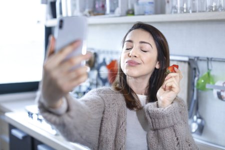 Téléchargez les photos : Brunette fille prend un selfie avec son téléphone portable tout en mangeant une fraise dans la cuisine - en image libre de droit