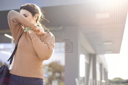 Foto de Chica con mascarilla y vestida casual estornuda y se repara con el brazo aislado en el centro comercial - Imagen libre de derechos