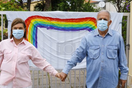 Photo for Elderly couple with surgical mask, casual dressed in front of a rainbow drawing on a gate outside their home, holding hands tenderly. - Royalty Free Image