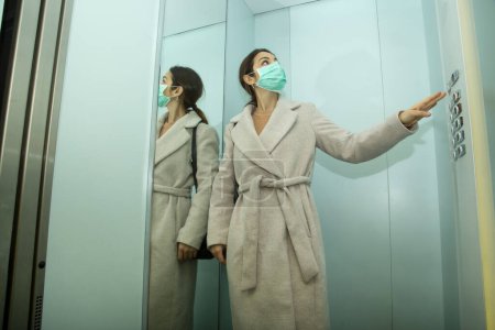 Téléchargez les photos : Brunette fille avec manteau prend l'ascenseur portant le masque protecteur - en image libre de droit