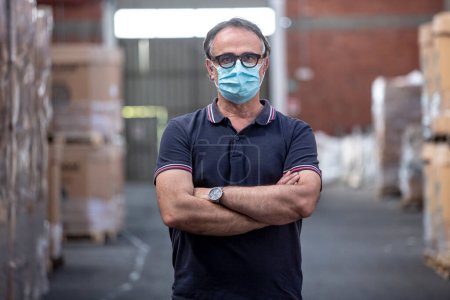 Photo for Man with black glasses and protective mask crosses his arms in the warehouse where he works - Royalty Free Image