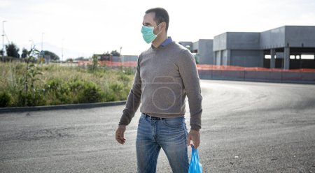 Téléchargez les photos : Casual habillé homme avec masque protecteur et sac à provisions traverse la rue - en image libre de droit