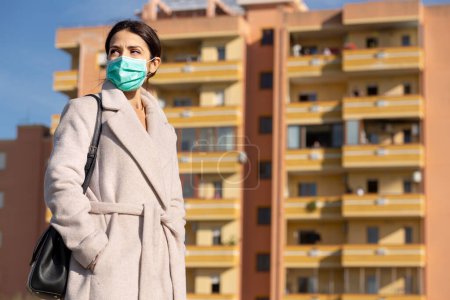 Photo for Woman in coat and mask looks serious, in the background a suburban building - Royalty Free Image