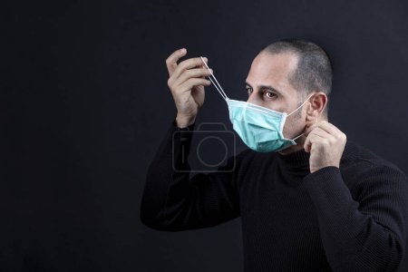 Foto de Hombre con el pelo afeitado usando una máscara quirúrgica, suéter negro, se quita la máscara, aislado sobre un fondo negro - Imagen libre de derechos