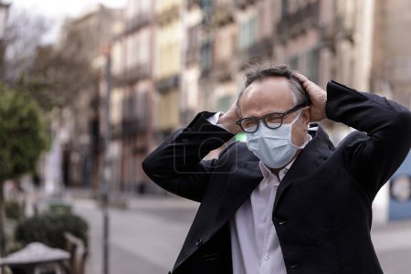 Photo for Portrait of middle-aged man in jacket with protective face mask, standing on a sidewalk with public greenery behind - Royalty Free Image