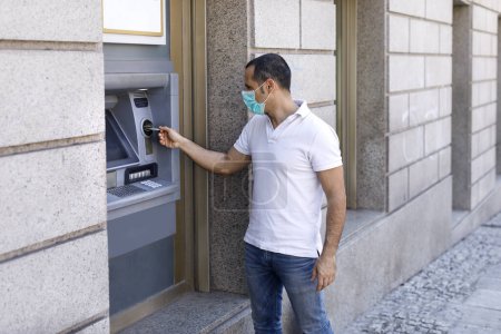 Photo for Casual dressed man with face mask withdraws money from an ATM on the street - Royalty Free Image