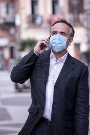 Photo for Man dressed in suit with protective face mask uses his cellphone in an urban context - Royalty Free Image