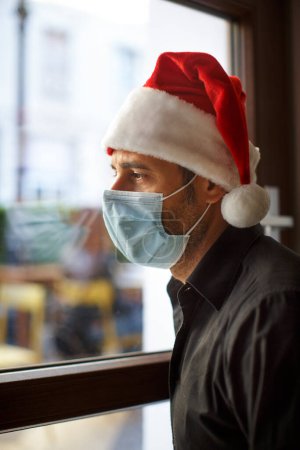 Photo for Man in Santa hat and surgical mask, looks sad outside the house through a window pane - Royalty Free Image