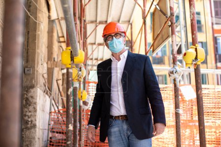 Téléchargez les photos : Ingénieur avec casque, veste élégante et masque est debout sous un échafaudage dans le centre-ville - en image libre de droit