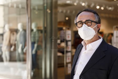 Photo for Man in jacket and surgical face mask is standing still in front of a clothing store in the city center - Royalty Free Image