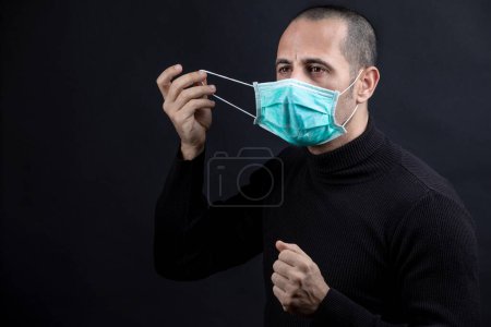 Photo for Man with shaved hair wearing a surgical mask, black sweater, takes off the mask, isolated on a black background - Royalty Free Image