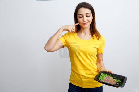 Foto de Mujer joven con cabello liso, lleva camiseta amarilla y mira el segundo plato de carne y guisantes con sabor, aislado sobre fondo blanco - Imagen libre de derechos