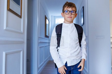 Téléchargez les photos : Blond petit garçon dans des lunettes et sac à dos est debout dans le couloir à la maison - en image libre de droit