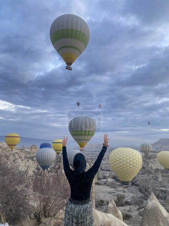 Téléchargez les photos : Femme aux cheveux bruns avec chemise noire et jupe fleurie levant les bras en signe d'exultation, en arrière-plan les montgolfières en Cappadoce en Turquie - en image libre de droit