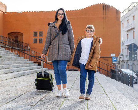 Téléchargez les photos : Une mère aux cheveux noirs accompagne son fils à l'école tout en tirant le sac à dos sur roues - en image libre de droit