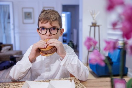 Foto de Niño rubio en vasos come una hamburguesa sentada en la mesa en casa - Imagen libre de derechos