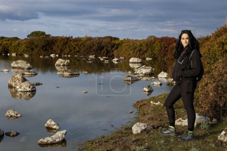 Foto de Mujer joven con pelo negro en abrigo negro de pie en la costa con rocas cerca del lago en el día de otoño - Imagen libre de derechos