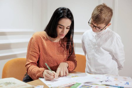 Photo for A dark-haired woman helps a blond boy with his homework sitting in an elegant room - Royalty Free Image