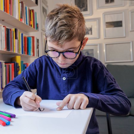 Téléchargez les photos : Blond garçon à lunettes et un polo bleu fait ses devoirs dans sa chambre - en image libre de droit