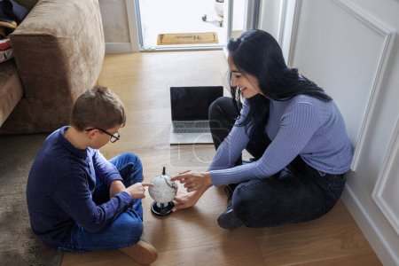 Foto de Chica adulta con el pelo oscuro muestra a un niño pequeño un globo mientras están sentados en el suelo en casa - Imagen libre de derechos