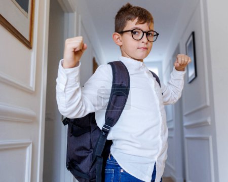 Téléchargez les photos : Enfant blond dans des lunettes et sac à dos debout dans le couloir à la maison montrant les muscles tout en jouant - en image libre de droit