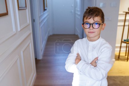 Téléchargez les photos : Petit garçon blond en lunettes et chemise blanche debout dans le couloir à la maison, croise les bras et semble sérieux - en image libre de droit