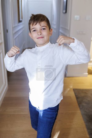 Téléchargez les photos : Enfant blond dans la chemise blanche debout dans le couloir à la maison, montrant les muscles tout en jouant - en image libre de droit