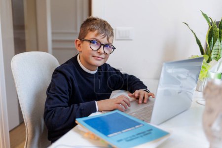 Téléchargez les photos : Garçon blond avec des lunettes, habillé en bleu et assis devant un bureau tout en travaillant sur l'ordinateur - en image libre de droit