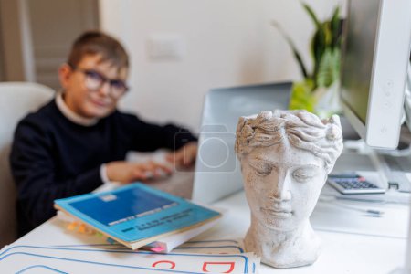 Téléchargez les photos : Garçon blond avec des lunettes, vêtu de bleu et assis devant un ordinateur tandis qu'une sculpture grecque se démarque au premier plan - en image libre de droit