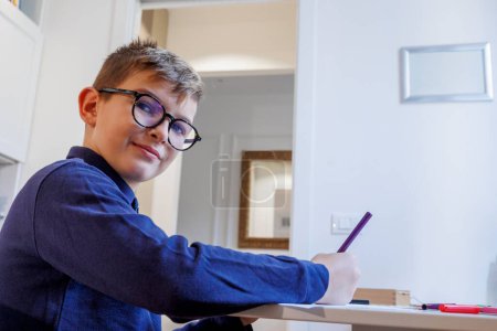 Foto de Niño rubio en gafas y una camisa de polo azul hace su tarea en su habitación - Imagen libre de derechos
