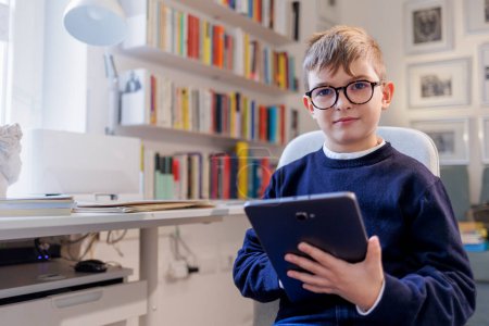 Téléchargez les photos : Garçon blond avec des lunettes, vêtu de bleu et assis devant un bureau, en utilisant une tablette à l'intérieur d'une étude avec une bibliothèque en arrière-plan. - en image libre de droit