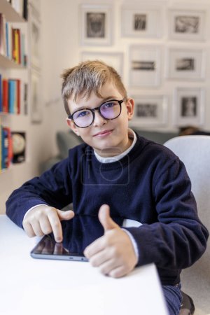 Téléchargez les photos : Garçon blond avec des lunettes, vêtu de bleu et assis devant un bureau, en utilisant une tablette à l'intérieur d'une étude avec une bibliothèque en arrière-plan. - en image libre de droit
