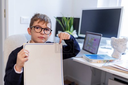 Foto de Niño rubio con gafas, vestido de azul y sentado frente a un escritorio, muestra un tablero con las letras del alfabeto - hay computadoras y monitores en el escritorio. - Imagen libre de derechos