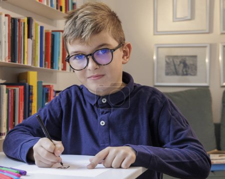 Téléchargez les photos : Blond garçon à lunettes et un polo bleu fait ses devoirs dans sa chambre - en image libre de droit